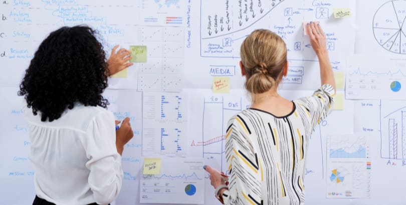 Two women analyzing data on a whiteboard covered with charts, graphs, and sticky notes.
