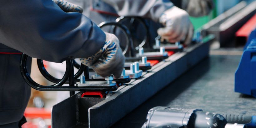 Workers assembling metal components on a production line. The image shows gloved hands fastening U-shaped metal brackets onto a flat base using bolts and washers, with tools visible on the workbench.
